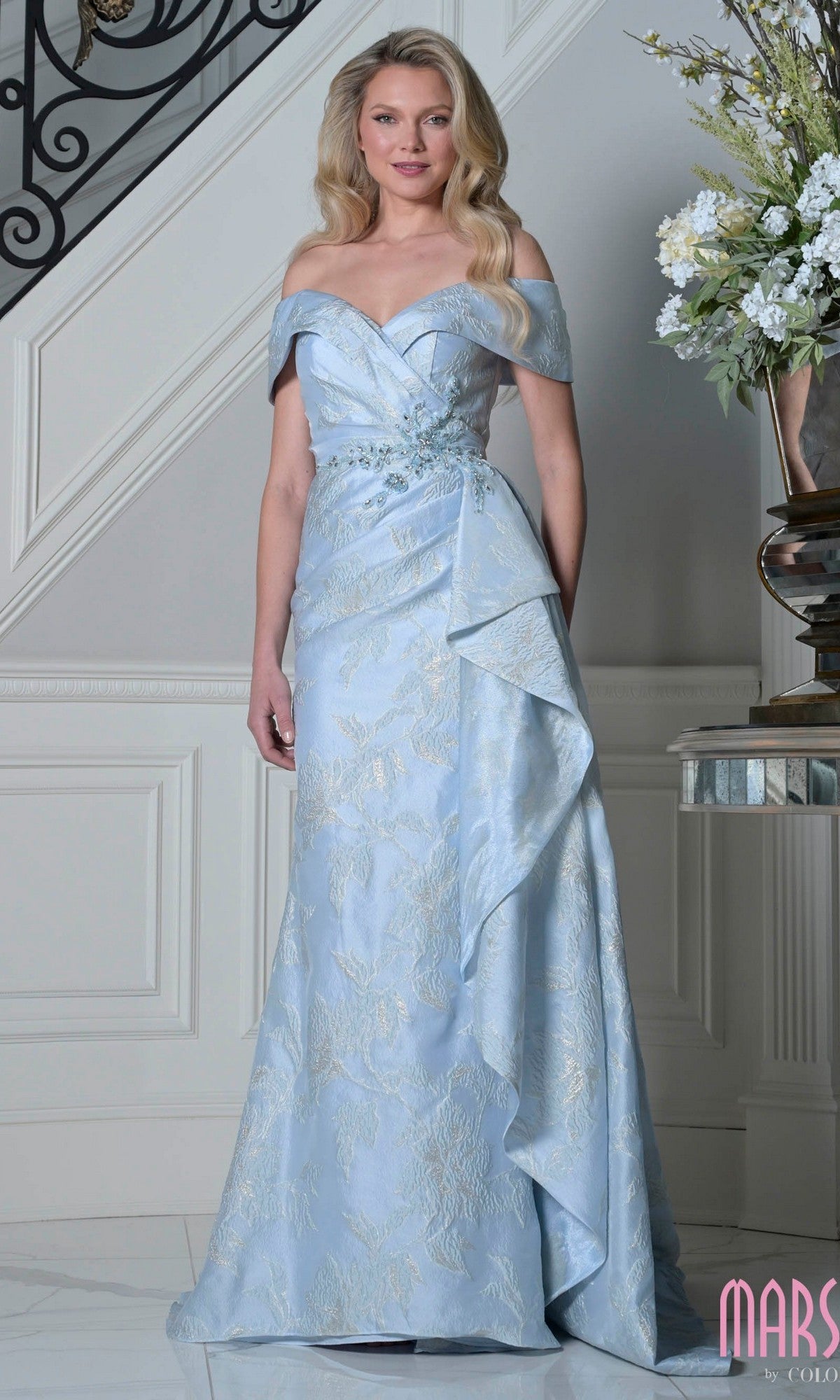 A woman in the Marsoni MV1401 long formal dress in powder blue, featuring off-the-shoulder style, floral patterns, and side draping, stands indoors near a white staircase and floral arrangement.